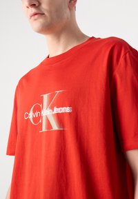 Crvena pamučna majica s istaknutim bijelim i srebrnim logom Calvin Klein Jeans, okrugli izrez, kratki rukavi i opušteni kroj.