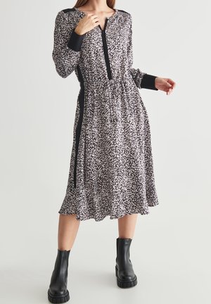 Nainen pukeutuneena pitkähihaiseen, mustavalkoiseen, kuvioituun polvipituiseen mekkoon, jossa on mustat sivupaneelit, ja mustiin nilkkureihin, seisomassa yksivärisen taustan edessä.