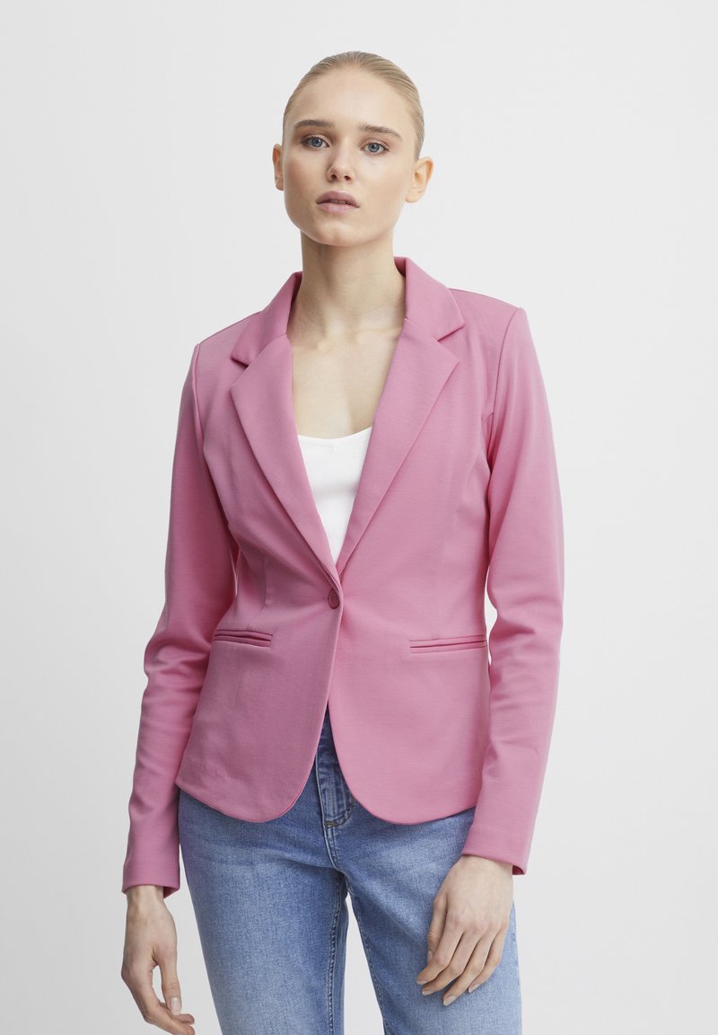 Lichtroze blazer met notch-revers, enkele knoopsluiting, twee zijzakken en een gladde textuur, gedragen over een witte top en blauwe jeans.