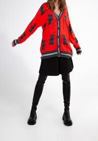Cárdigan de punto rojo con patrón de osito de peluche negro, puños acanalados y acentos grises a rayas. Combinado con una falda negra y botas negras brillantes.
