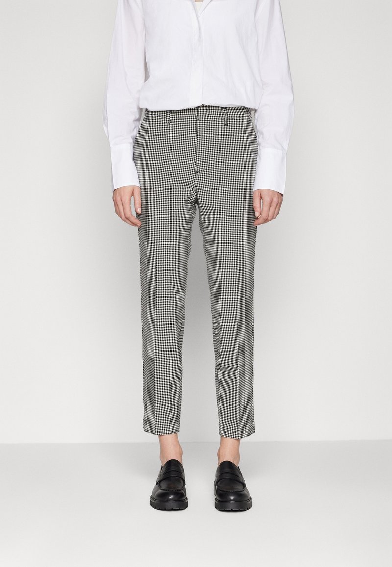 Personne portant un pantalon slim à motif pied-de-poule noir et blanc, une chemise blanche à manches longues boutonnée et des mocassins noirs, debout.