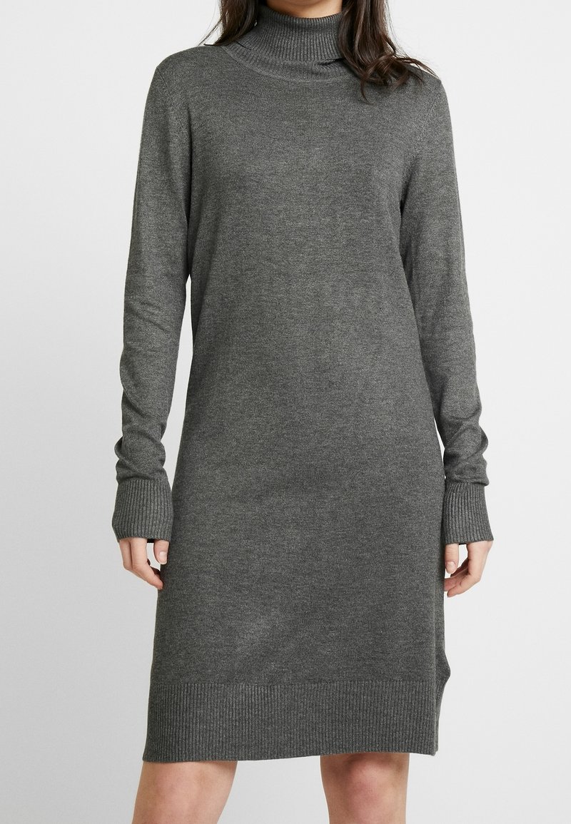 Femme portant une robe pull col roulé gris foncé à manches longues, longueur au genou, avec des poignets et un ourlet côtelés.