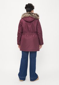Casaco acolchoado cor vinho com capuz de pelúcia sintética, cintura ajustada e mangas longas. Usado com calças de ganga flare e botas cor de pele.