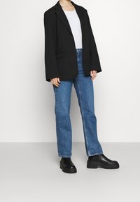 Blazer negro extragrande, top blanco ajustado, jeans de pierna recta azul y botas de tobillo negras gruesas, todo en un fondo claro.