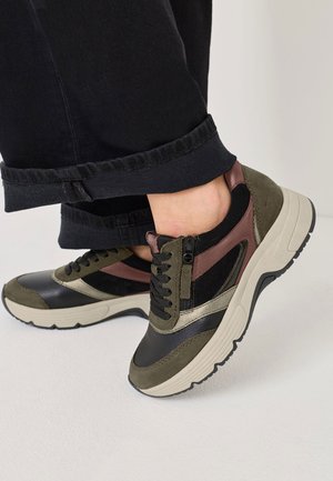 Fødder iført chunky sneakers med sorte, olivengrønne, mørkerøde og beige detaljer, parret med opsmurte sorte bukser på et hvidt gulv.