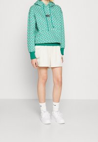 Green patterned Nike hoodie with a front pocket, paired with beige shorts. Model wears white socks and shoes, creating a casual look.