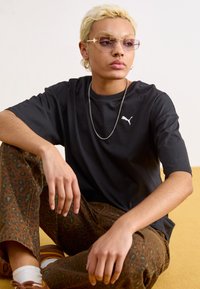 Tricou negru din bumbac cu logo-ul PUMA, asortat cu pantaloni cu imprimeu leopard în nuanță de maro. Accentele includ ochelari de soare aurii și un colier din lanț argintiu.