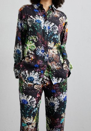 Kvinde iført sort skjorte med knapper og bukser med farverigt, abstrakt blomsterprint, hænderne i lommerne, foran ensartet grå baggrund.