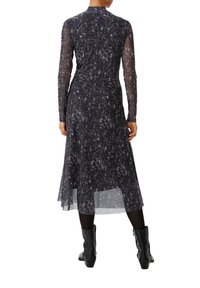 Schwarzes Langarmkleid mit transparenter Überlage, präsentiert ein weißes abstraktes Muster, tailliert mit elastischem Bund und Midi-Länge. Trägt schwarze Stiefel.