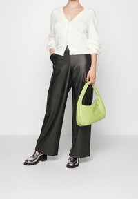 Person i vit, fluffig, knappad kavaj, svarta vida byxor, metalliskt silverfärgade loafers, som håller en ljusgrön handväska vid sin sida.