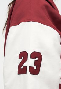 Krupni plan rukava bijele i bordo jakne s brojem 23 ušivenim bordo koncem na bijelom materijalu.