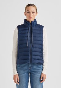 Marineblaue gesteppte Daunenweste mit hohem Kragen, horizontalem Steppmuster, Reißverschluss vorne, Kordelzügen und seitlichen Taschen. Wird über einem leichten Pullover getragen.