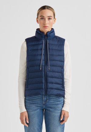 Marineblaue gesteppte Daunenweste mit hohem Kragen, horizontalem Steppmuster, Reißverschluss vorne, Kordelzügen und seitlichen Taschen. Wird über einem leichten Pullover getragen.