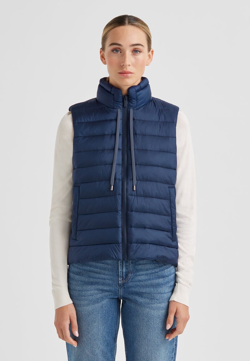 Marineblaue gesteppte Daunenweste mit hohem Kragen, horizontalem Steppmuster, Reißverschluss vorne, Kordelzügen und seitlichen Taschen. Wird über einem leichten Pullover getragen.