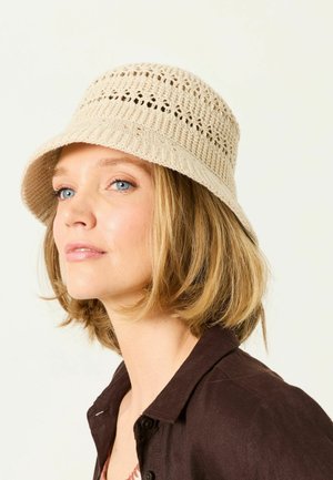Kobieta o jasnobrązowych włosach nosi beżowy szydełkowany kapelusz typu bucket oraz ciemnobrązową koszulę z kołnierzykiem, patrząc lekko na bok.
