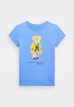 T-shirt enfant bleu clair à manches courtes avec un motif d'ours en peluche portant une veste jaune, un short à carreaux et des baskets.