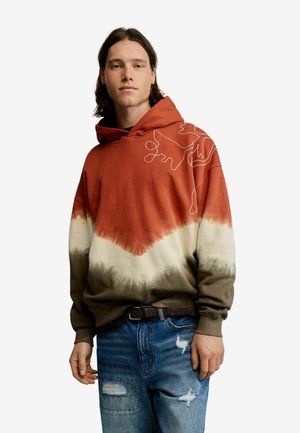 Vállig érő hajú fiatal férfi, aki piros, fehér és barna batikolt kapucnis pulóvert visel, hímzett absztrakt mintával és szakadt kék farmerral.