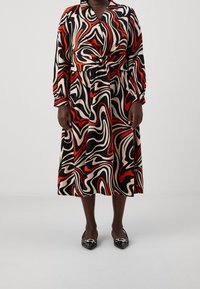 Vestido de manga larga, hasta la rodilla, con un patrón en espiral de rojo, negro y crema, con cintura atada y cuello abotonado. Combinado con bailarinas negras.