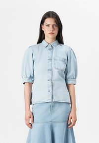 Model wearing a light blue denim shirt with puffed sleeves and a chest pocket, paired with a matching denim skirt, against a white background.