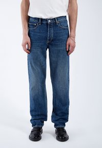 Jeans in denim blu con una vestibilità rilassata, gambe affusolate, cinque tasche e hardware in metallo. Indossati con una t-shirt bianca semplice e scarpe nere.