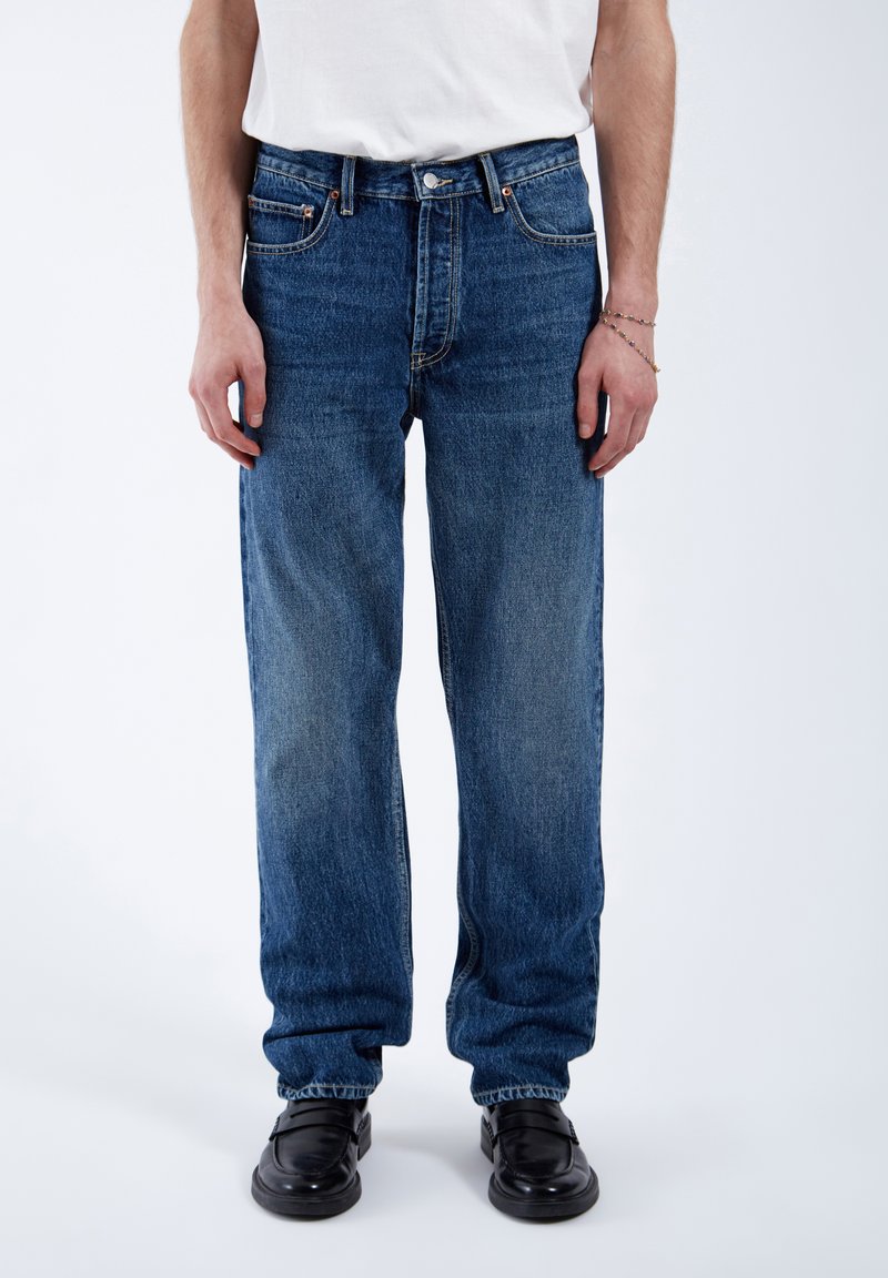 Jeans in denim blu con una vestibilità rilassata, gambe affusolate, cinque tasche e hardware in metallo. Indossati con una t-shirt bianca semplice e scarpe nere.