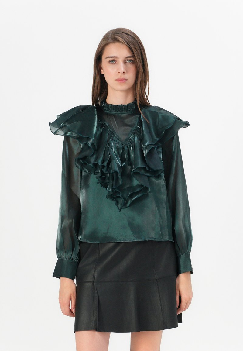 Groene satijnen blouse met grote rimpelkragen en mouwen, gecombineerd met een zwarte leren rok. Heeft een peplum-silhouet en een gestructureerde stof.