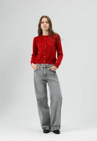 Cardigan rouge en maille avec encolure ronde et fermeture boutonnée à l'avant, associé à un jean gris à taille haute et jambes larges. Texture lisse, coupe décontractée.