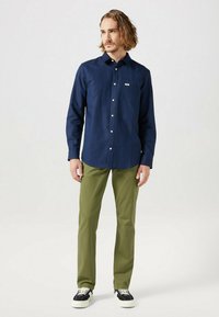 Camicia blu navy con bottoni in tessuto leggero, dotata di taschino sul petto, abbinata a pantaloni slim-fit verde oliva e sneakers nere con suola bianca.