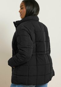 Doudoune noire matelassée avec col haut, présentant une couture en grille et une coupe décontractée. Texture douce et poches latérales visibles.