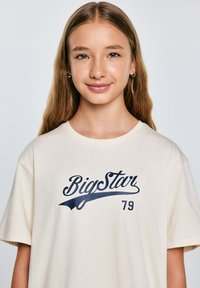 Beige kokvilnas T-krekls ar tumši zilā krāsā „BigStar” logotipu un skaitli „79” izteiksmīgā sportiskā fontā. Īsas piedurknes, relaksēta piegriezuma.