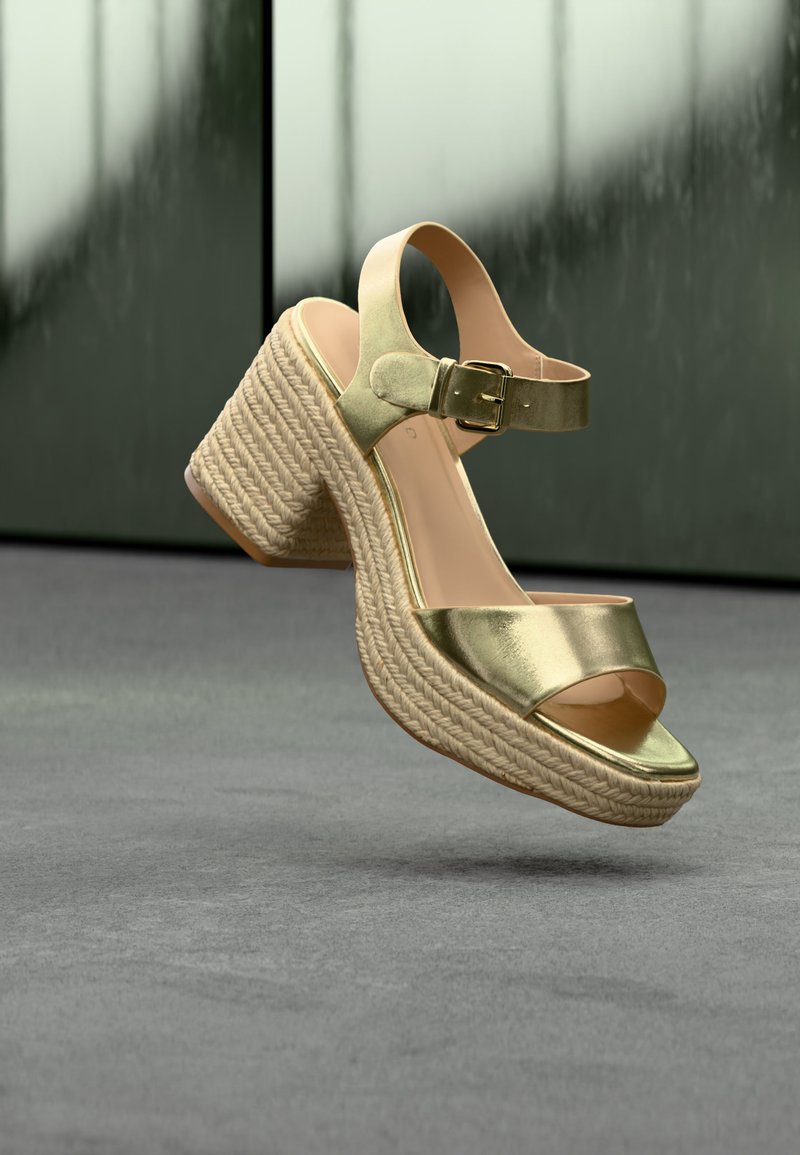 Sandale compensée métallique dorée à bout ouvert avec bride à la cheville et boucle, semelle plateforme en corde tressée, flottant au-dessus d'un sol gris.
