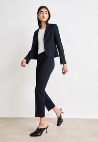 Blazer sartoriale navy con due bottoni, abbinato a pantaloni navy slim fit e mule nere con tacco aperto. Tessuto liscio con dettagli minimi.