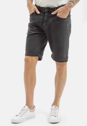 Mężczyzna w czarnych podwiniętych szortach dżinsowych i białych tenisówkach, z rękami w kieszeniach, stojący na jednolitym tle.