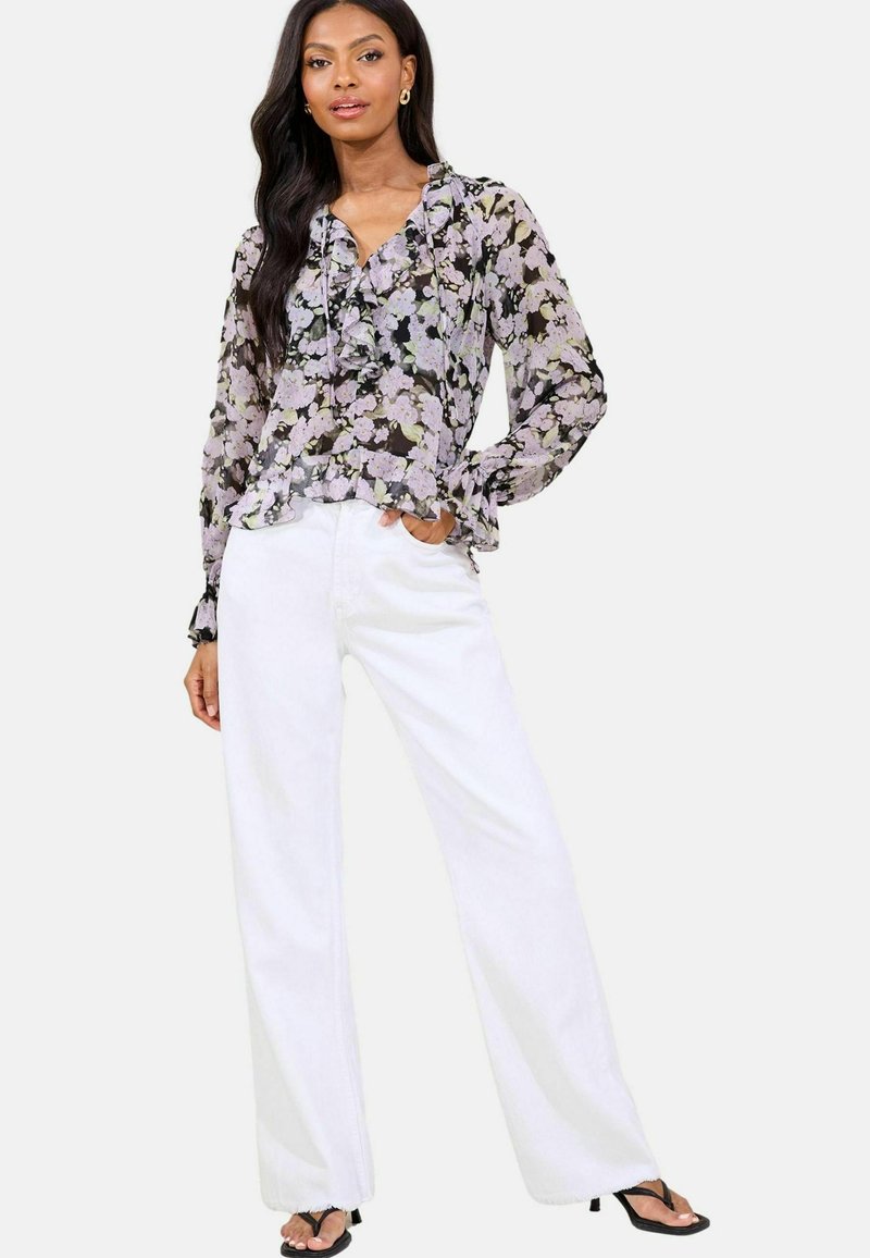 Blumenbluse mit Rüschen in Schwarz, Lavendel und Grün auf transparentem Stoff; kombiniert mit weißen hochgeschnittenen Weitenhosen und schwarzen Sandalen.