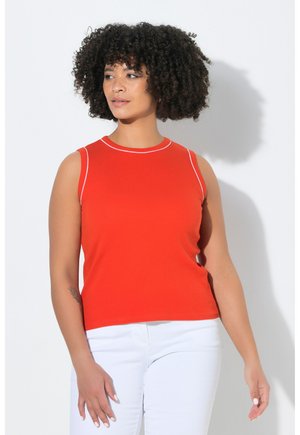 Femme aux cheveux bouclés portant un haut rouge sans manches avec un liseré blanc et un pantalon blanc, debout devant un fond blanc.