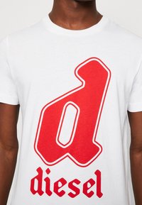 Vit bomull t-shirt med en stor röd stiliserad "d" med röd kontur och varumärket "diesel" i fet svart typografi nedanför.