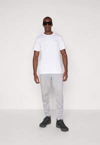 Hvid kortærmet t-shirt med logo, lysegrå sweatpants og sorte sportssko. Modellen har mørke solbriller på. Simpelt, afslappet outfit.