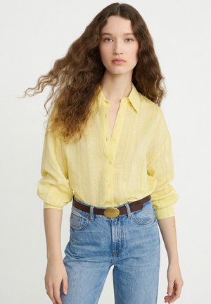 Jeune femme aux longs cheveux ondulés portant un chemisier jaune boutonné rentré dans un jean bleu taille haute avec une ceinture marron.