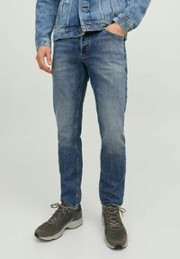 Jeans de denim azul claro, ajustados y con un acabado desgastado. Presenta cinco bolsillos y costuras naranjas. Combinados con zapatillas deportivas grises.