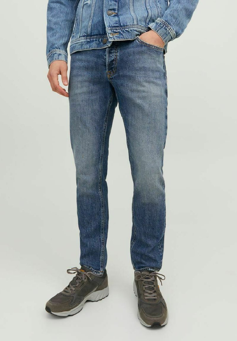 Jeans de denim azul claro, ajustados y con un acabado desgastado. Presenta cinco bolsillos y costuras naranjas. Combinados con zapatillas deportivas grises.