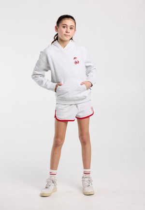 Junges Mädchen, das mit den Händen in den Taschen steht, trägt einen hellgrauen Hoodie mit roter Kirschen-Grafik, passende Shorts mit rotem Rand und weiße Turnschuhe.
