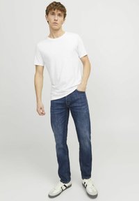Weißes T-Shirt mit kurzen Ärmeln, blaue gerade Jeans und weiße Sneakers mit schwarzen Streifen, stehend vor einem neutralen Hintergrund.
