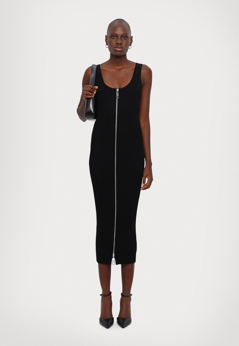 Femme en robe midi sans manches noire à côtes avec fermeture éclair frontale, tenant un sac à épaule noir, portant des boucles d'oreilles créoles et des talons noirs.