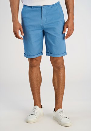 Blaue Baumwollshorts mit aufgerollten Säumen, versehen mit einem Reißverschluss und Knopfverschluss. Kombiniert mit weißen Turnschuhen ist die Textur glatt und entspannt.