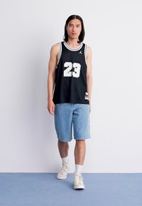 Zwarte basketbaltrui met nummer 23, witte accenten en denim shorts. Model draagt beige sneakers en witte sokken, staand op een blauwe vloer.
