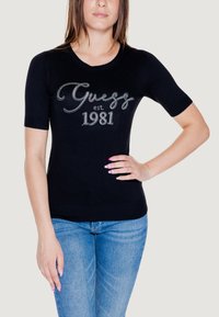 Maglione nero a maniche corte con scollatura rotonda, caratterizzato da una grafica in strass argento "Guess est. 1981" sul davanti. Texture del tessuto morbido.