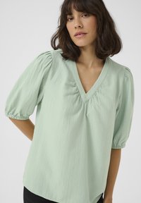 Blouse vert clair, texturée, avec un col en V et des manches bouffantes froncées. Le tissu présente un motif subtil et une coupe décontractée.