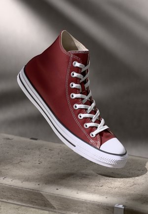 Zapatilla alta roja de lona, con cordones blancos, puntera de goma y ribete negro a lo largo de la suela. Superficie texturizada con detalles de costura.