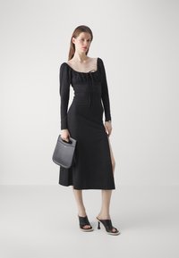 Black midi dress with puff sleeves, fitted bodice, and side slits. Paired with black heeled sandals and a small black handbag.