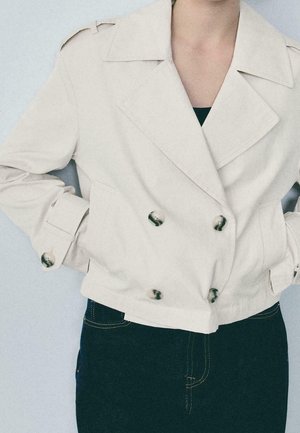 Persoon draagt een beige cropped double-breasted jas met grote knopen en zwarte broek, handen in de jaszakken.